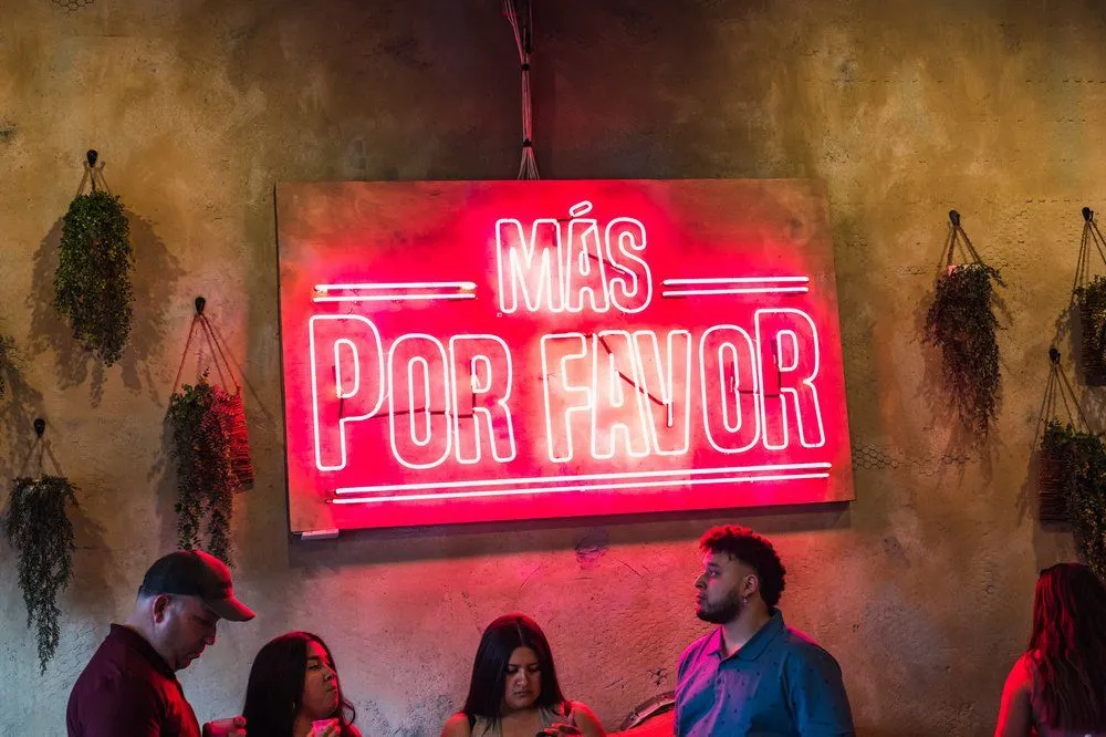 Interior of Más Por Favor in Las Vegas featuring neon signage and guests enjoying tacos and cream cheese churros in a vibrant, modern taqueria setting.